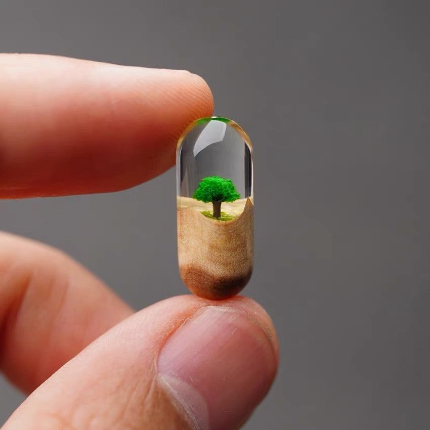 Pill with a miniature landscape including a tree held between fingers against a gray background