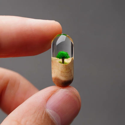 Pill with a miniature landscape including a tree held between fingers against a gray background