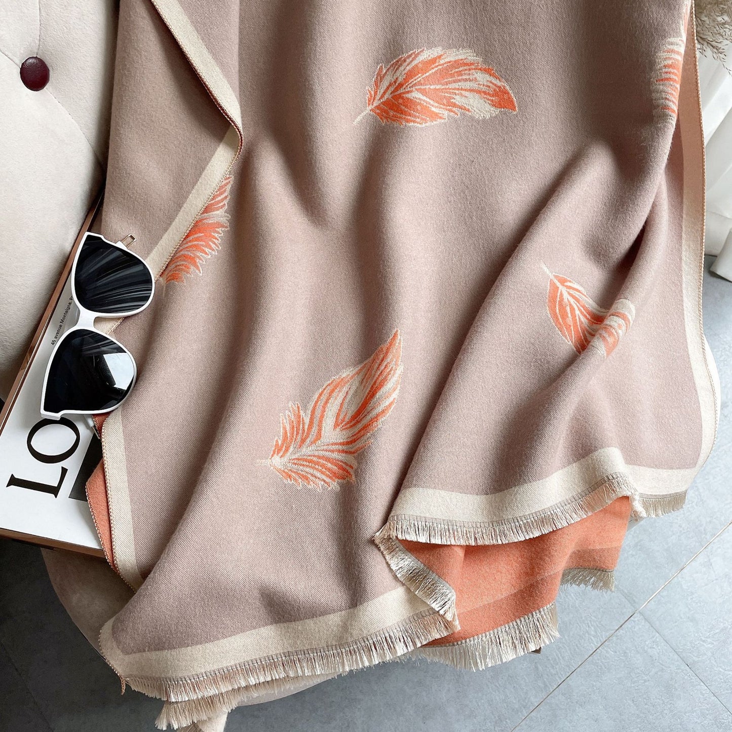 Beige scarf with feather patterns draped over a magazine and sunglasses on a light gray surface.